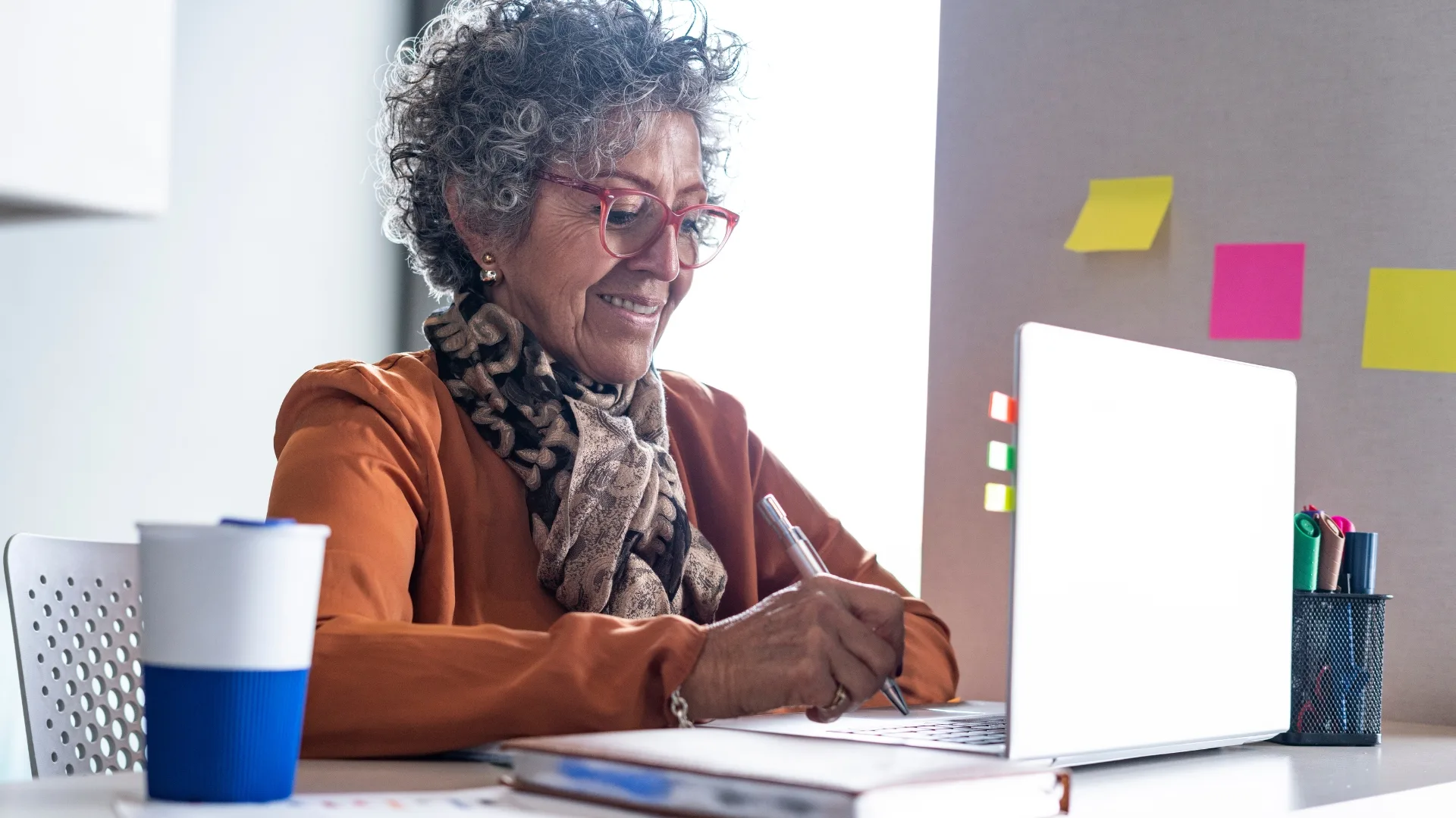 Older woman wearing an orange top and patterned scarf sits at a desk with a laptop.