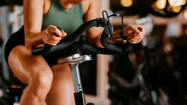 Woman in a green top and black shorts intently gripping the handlebars of a stationary exercise bike in a gym.