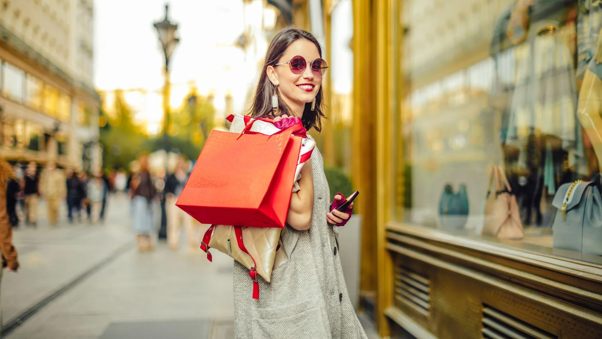 A stylish woman with sunglasses smiles over her shoulder while holding shopping bags and a phone, walking past storefronts on a busy street.
