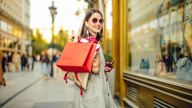 A stylish woman with sunglasses smiles over her shoulder while holding shopping bags and a phone, walking past storefronts on a busy street.