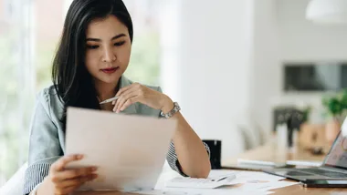 A woman wearing a blue shirt sits at a desk looking at a sheet of papers.