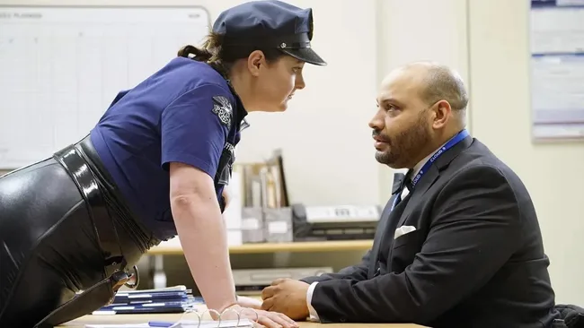 A woman in a police Halloween costume leans over a desk talking to a man in a suit.