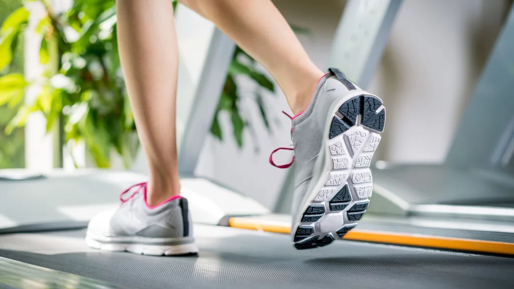 A close-up shot of a person's feet and legs while running or walking on a treadmill. The person is wearing grey and white athletic shoes with pink laces, showing the dark, textured sole of the shoe as the foot lifts off the belt.
