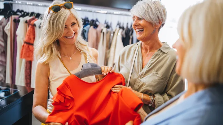 Two laughing, middle-aged women in a clothing store look at a bright red dress on a hanger, talking to a third person whose back is to the camera.