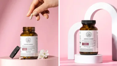 A two-panel image showing the Happy Mammoth Hormone Harmony supplement. On the left, a hand holds a capsule above an open bottle resting on a pink platform with a white flower. On the right, a bottle stands on a white pedestal with a pink background.