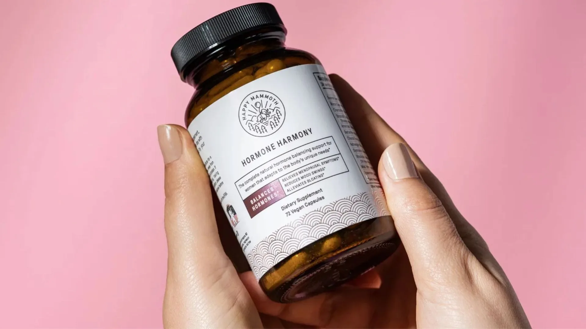 Close-up of a person holding a brown glass bottle of "Happy Mammoth Hormone Harmony" dietary supplements against a pink background.