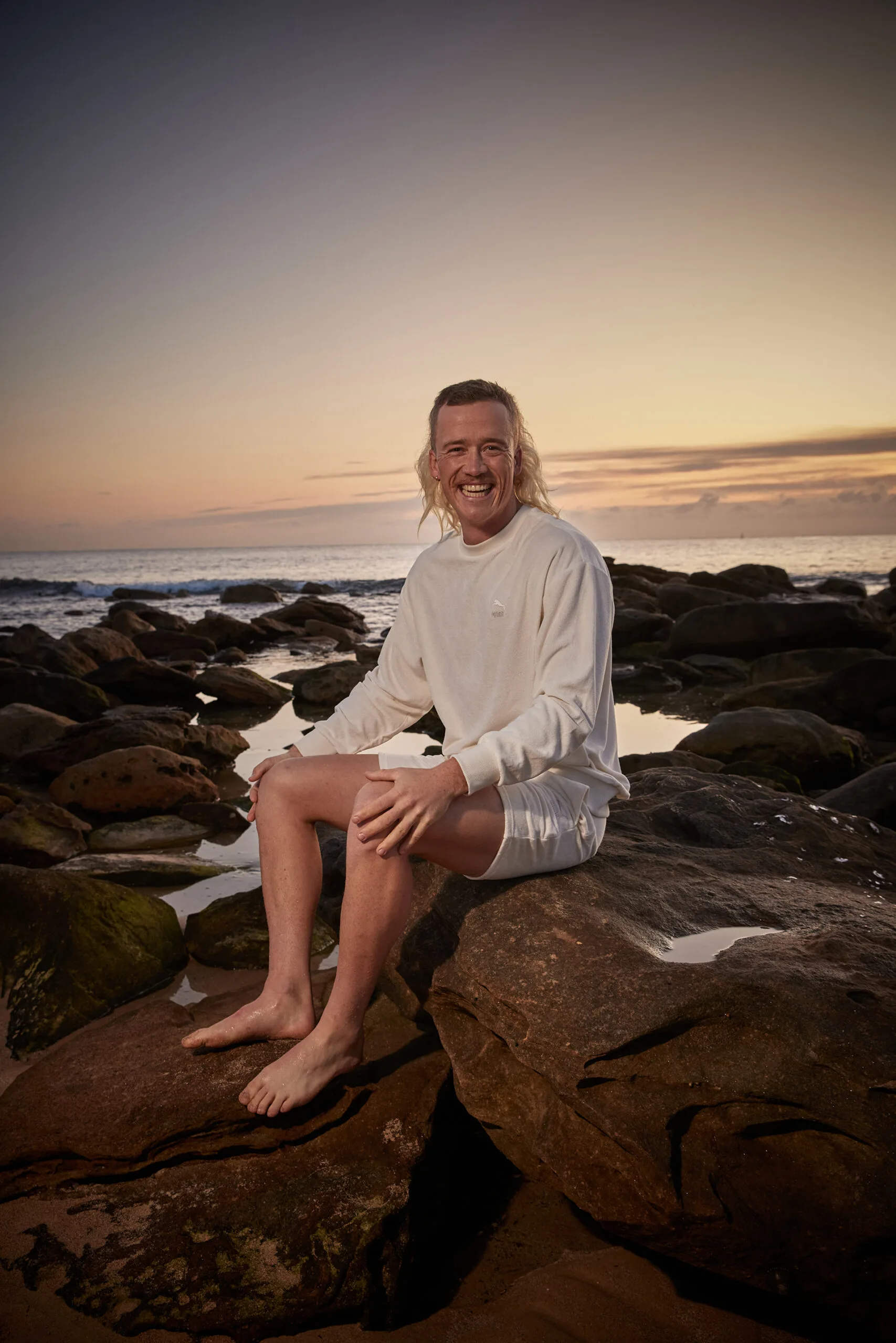 Nedd wears white boardshorts and top and sits amongst rockpools at a Sydney beach.