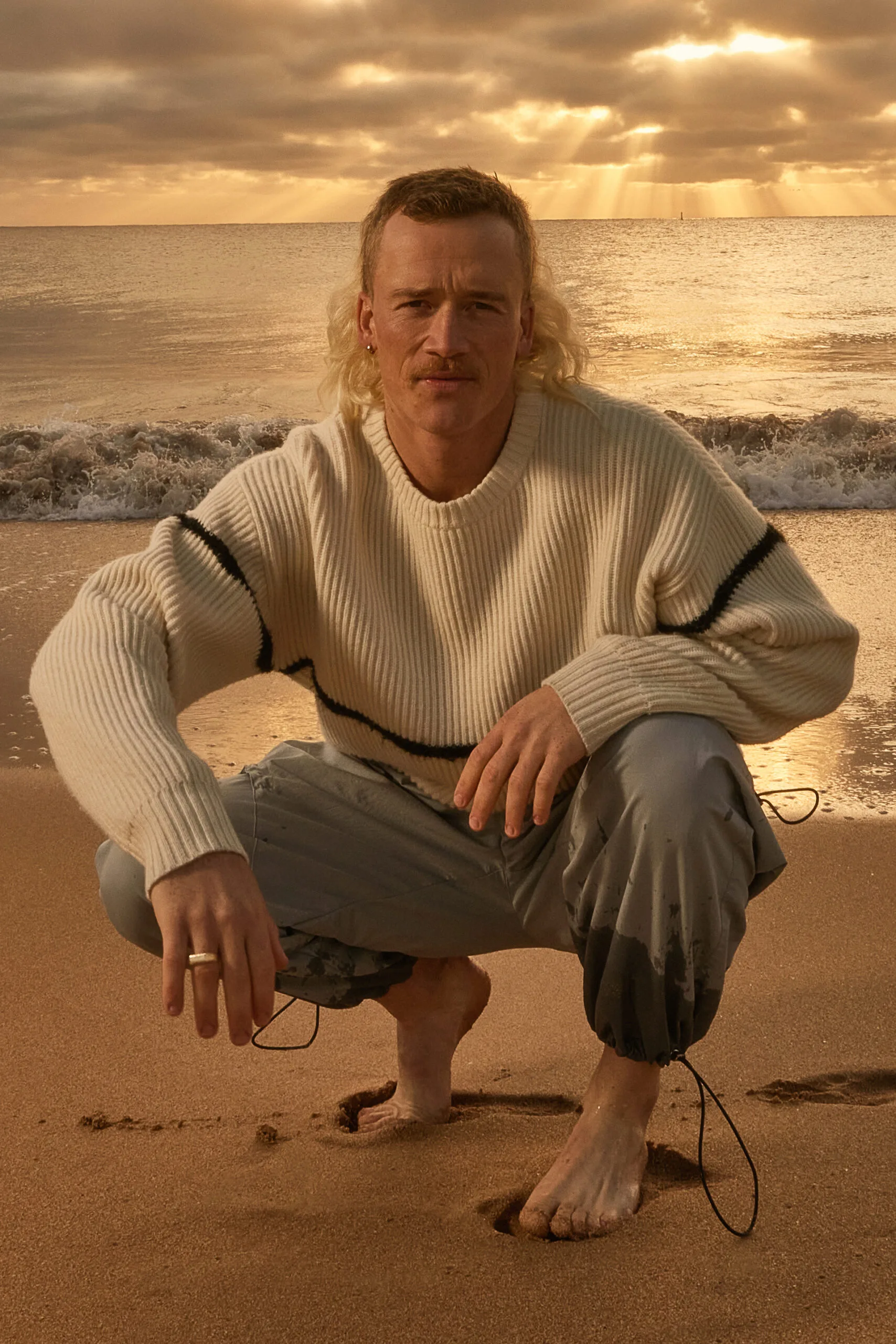 Nedd crouches on the sand at sunrise. Waves roll in behind him.