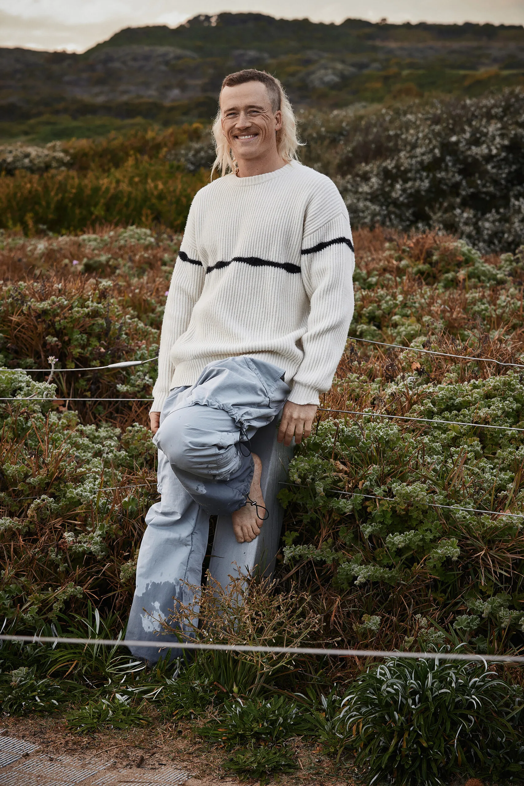 Ness leans against a fence post with coastal heath behind him.