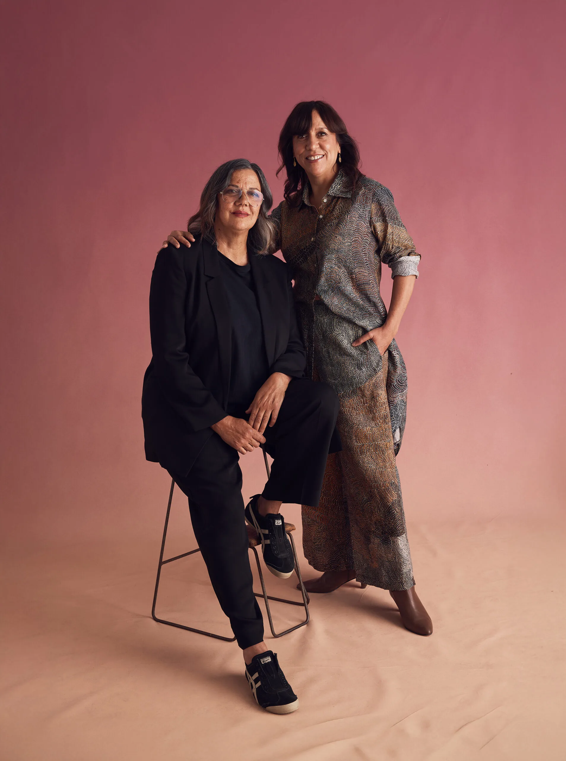 A photograph of Hetti Perkins seated, wearing black, and Rachel standing beside her with her arm on Hetti's shoulder. The background is pink and brown ombre shades.