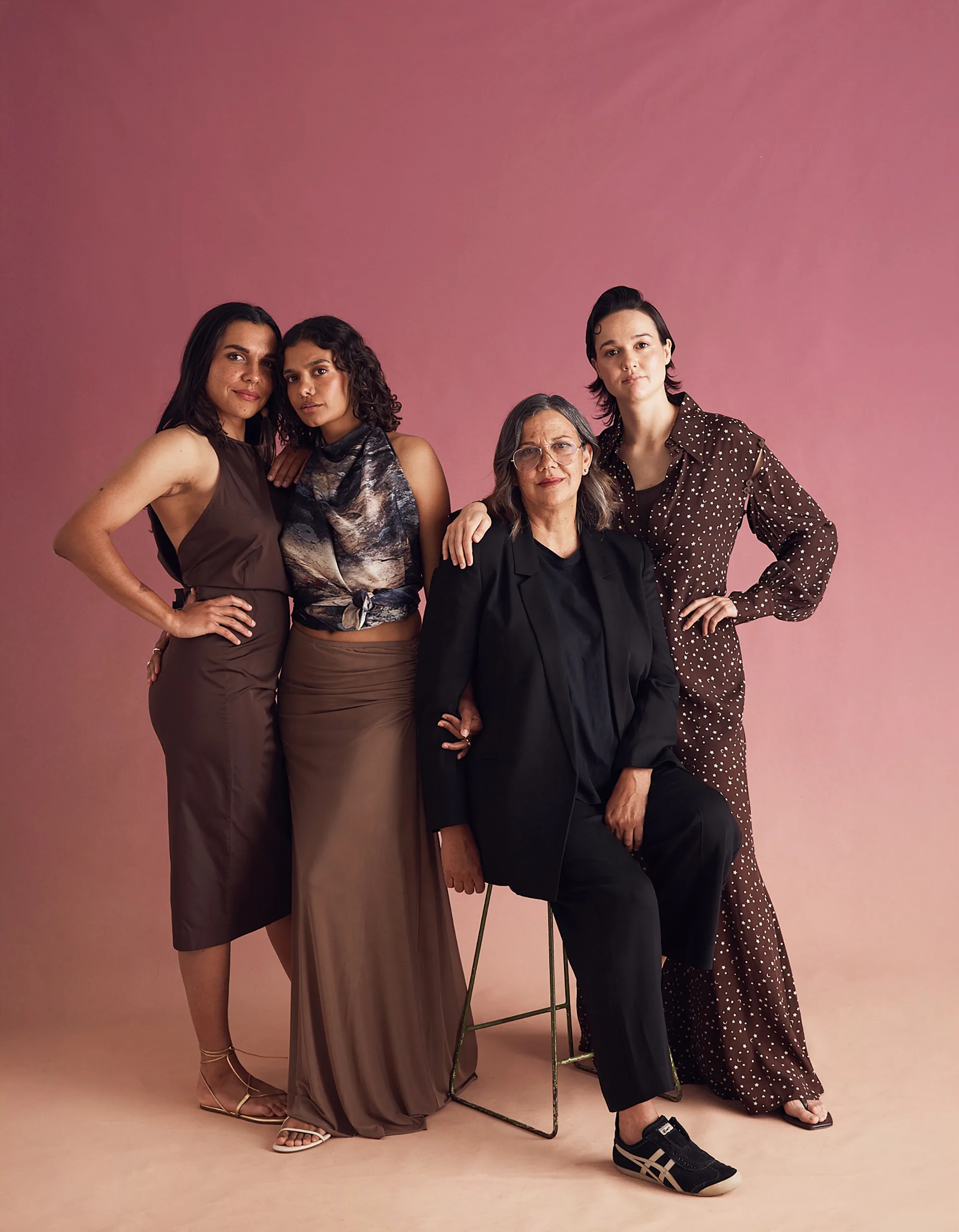 Hetti Perkins is seated and surrounded by her three daughthers. The photograph was taken in a studio with a pink and brown ombre background.