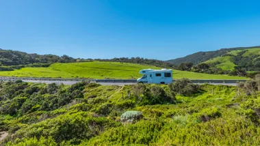 Campervan hire driving on a scenic road against a backdrop of lush green landscape and bright blue sky, perfect for an Australian road trip for over 50s.