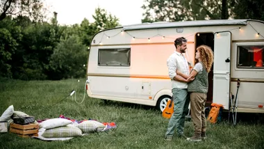 A smiling couple embracing outside a small campervan decorated with string lights, pitched in a grassy field.
