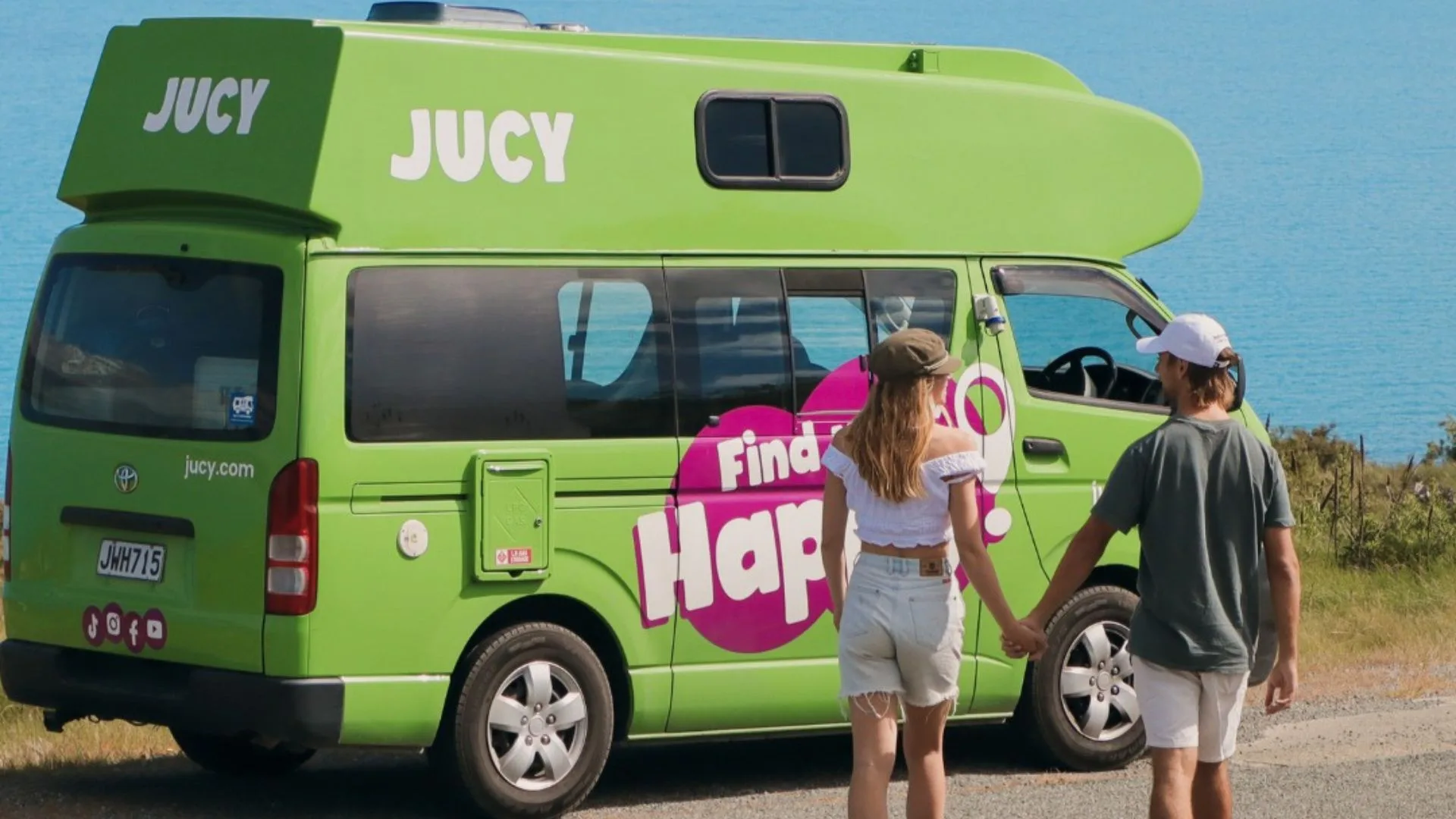 JUCY campervan hire - a man and woman holding hands walking toward a bright green campervan. The ocean is in the background.