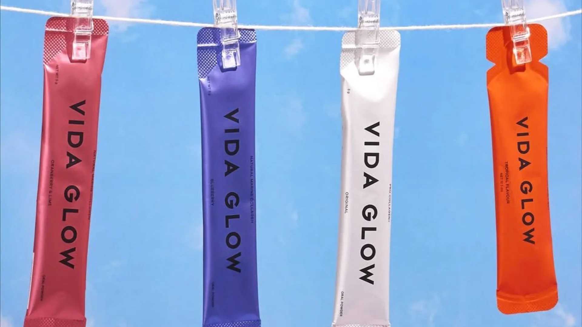 An outdoor product shot featuring four colourful Vida Glow sachets hanging from a clothesline against a bright blue sky. From left to right, the flavours shown are: Cranberry & Lime (pink), Blueberry (purple), Original Marine Collagen (white), and Tropical Flavour (orange).