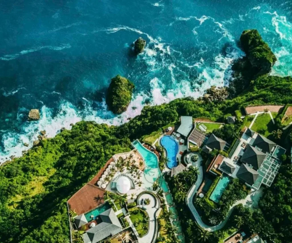 Aerial view of a luxury cliffside resort in Bali with turquoise ocean waves, lush greenery and multiple swimming pools.