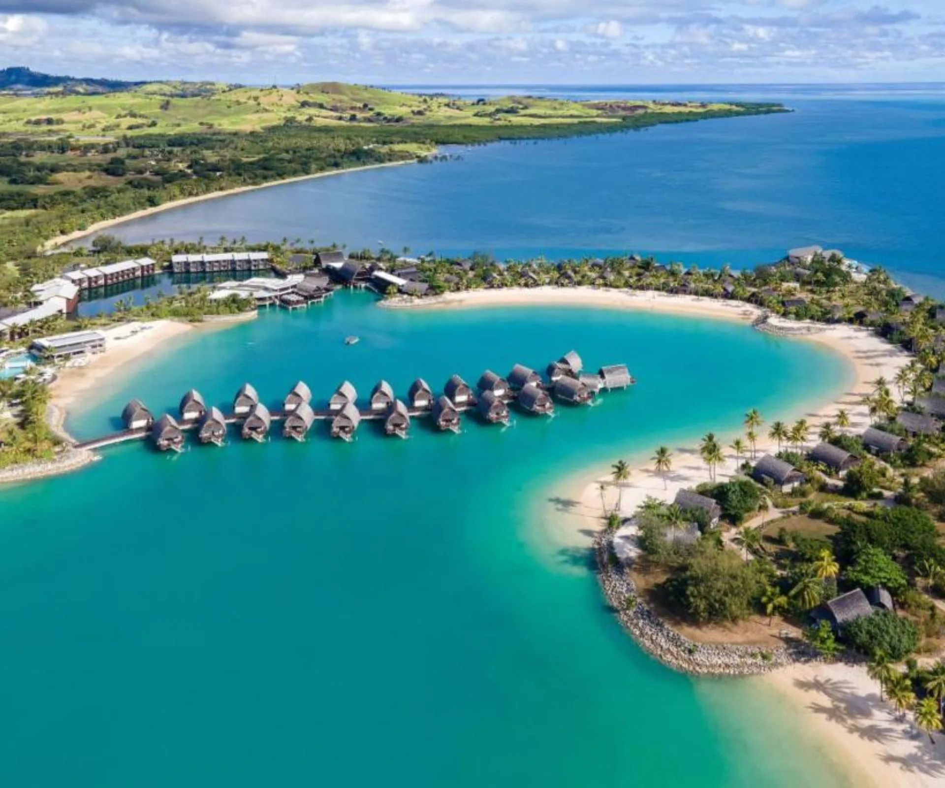 Aerial view of a luxury Fiji resort featuring a line of thatched-roof overwater bungalows sitting in a bright turquoise lagoon.