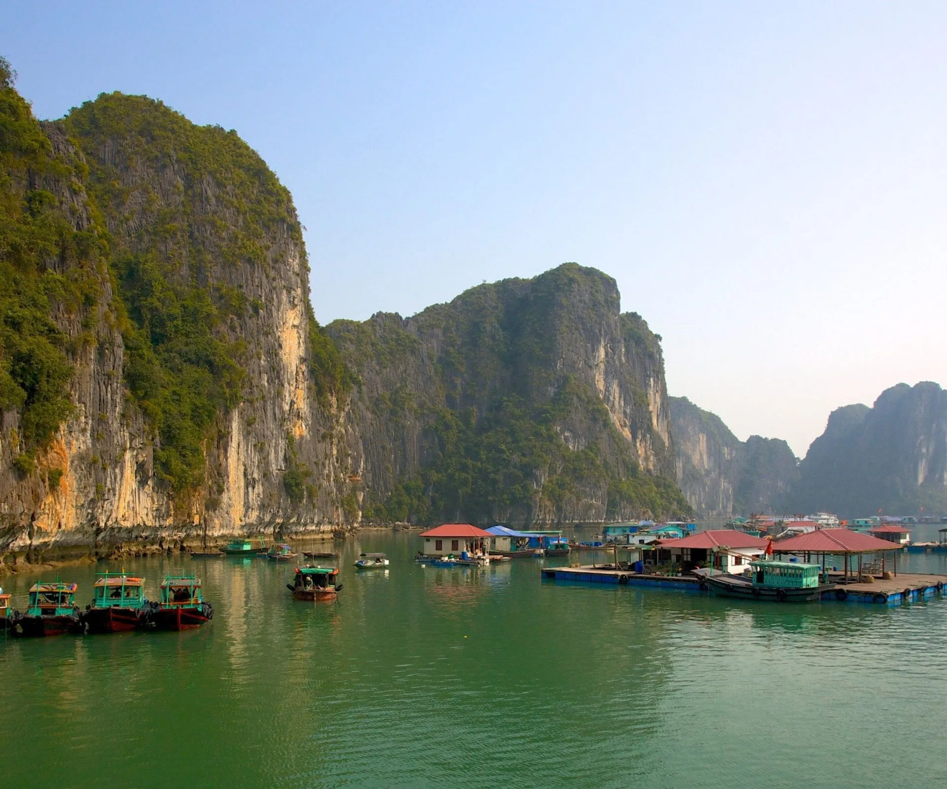 A floating village with red-roofed houses and small wooden boats nestled among the towering limestone karsts of Ha Long Bay, Vietnam.