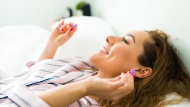A woman in silk pyjamas lying in bed, smiling as she inserts a purple earplug; part of a guide on the best earplugs for sleep in Australia.