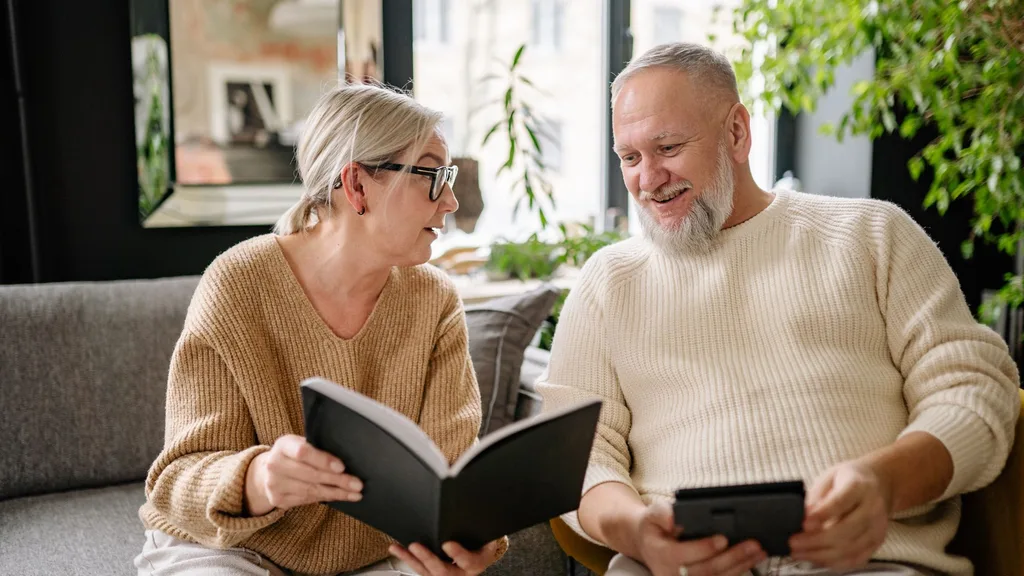 A woman and man on a couch talking. She holds a book and he has a kinidle.