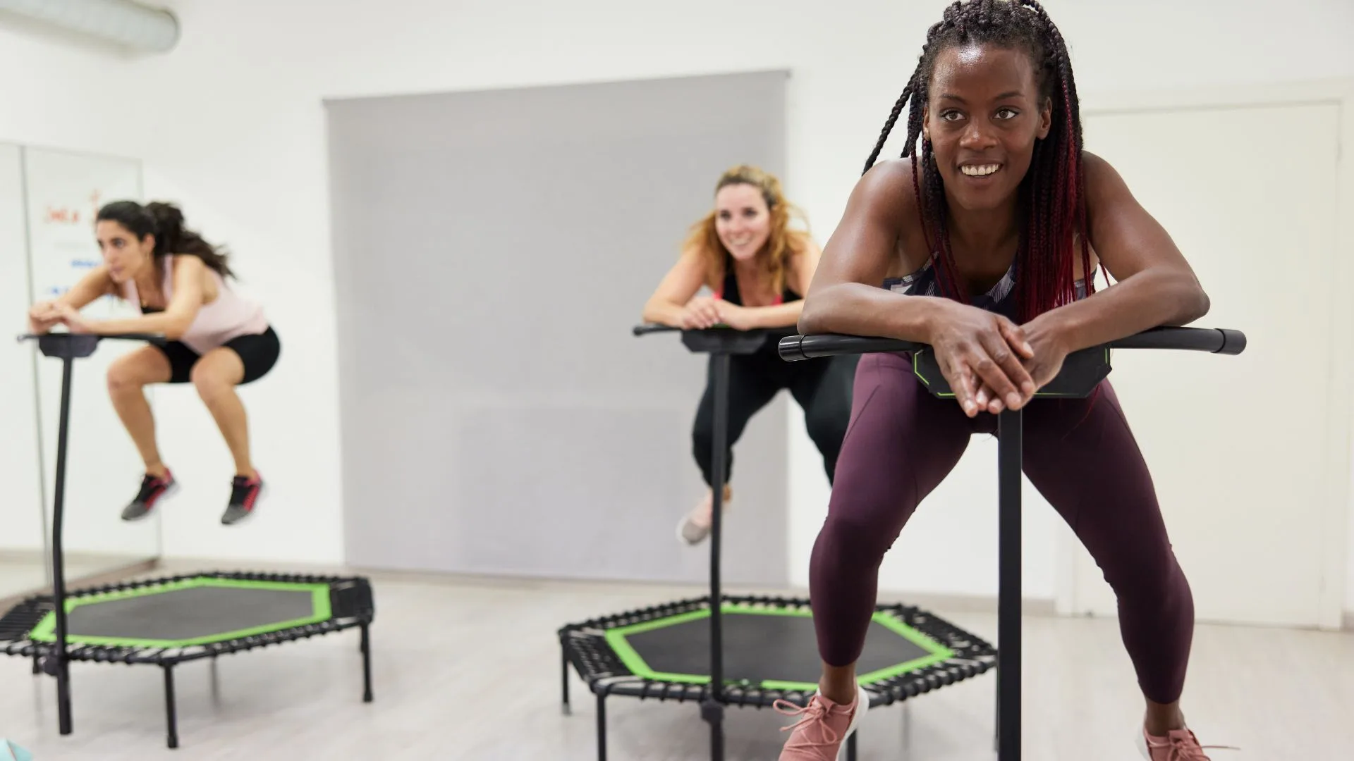 Three women participating in a rebounder fitness class in a bright, minimalist studio. In the foreground, a smiling woman with long braids leans against the stability bar of her hexagonal rebounder trampoline, while two other women are seen mid-workout in the background.