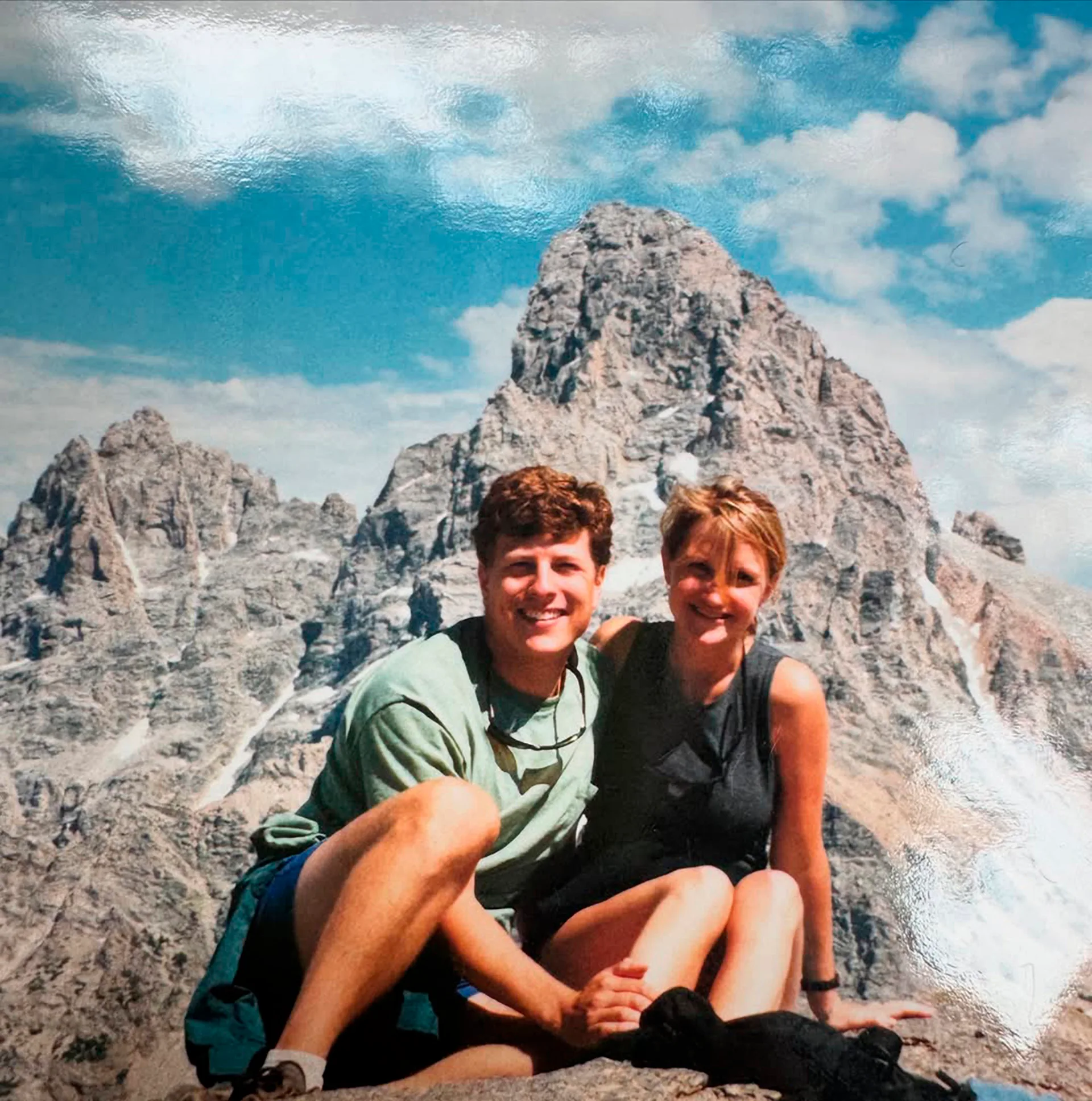 Chris and Mel, together on a hike, sit on a rocky outcrop with a mountainpeak behind them.