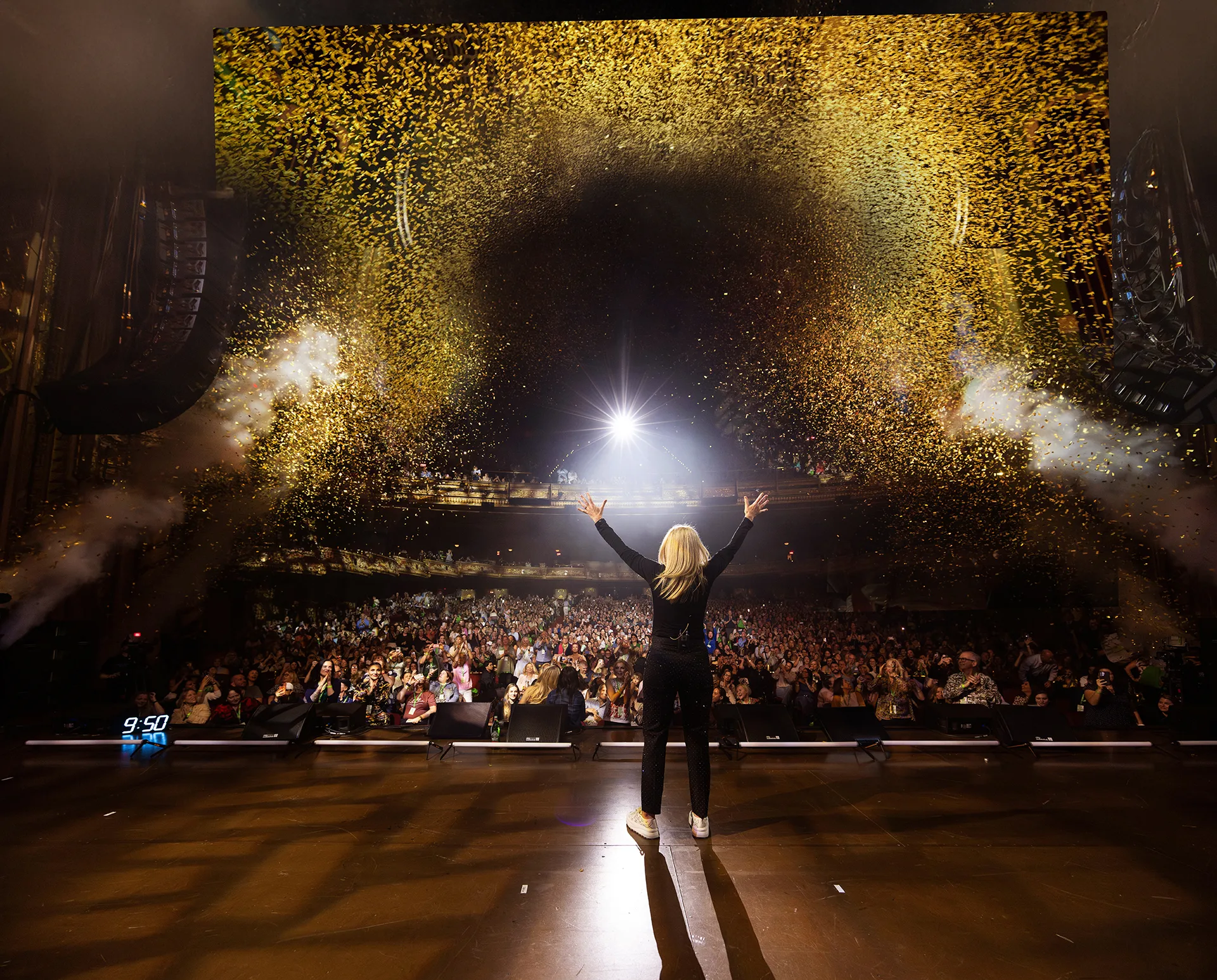 Mel, wearing a black pantsuit, seen from behind, raising her arms to the audience. The air is full of gold sparkles.