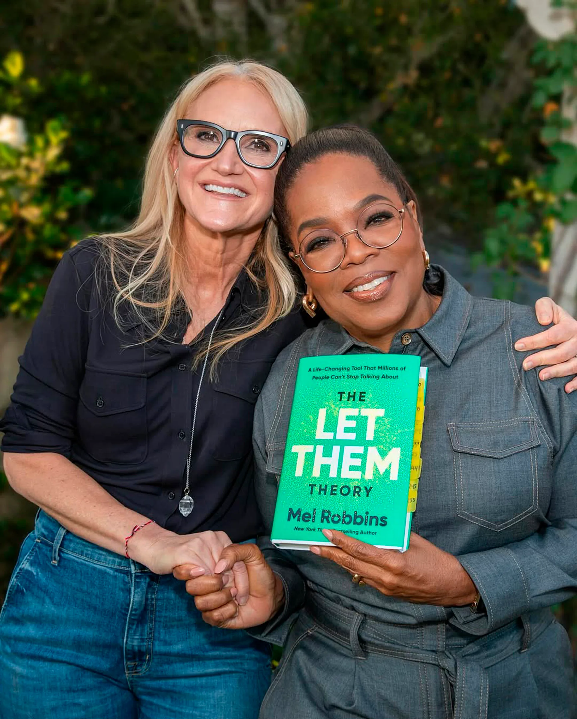 Oprah holds a copy of 'The Let Them Theory' which has a gren cover, as she stands with Mel, arm in arm.