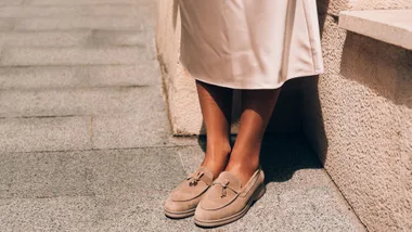 Close-up of a woman’s feet in light brown suede loafers with decorative gold charms and tassels. She is wearing a cream-coloured midi skirt against a sunlit stone wall.
