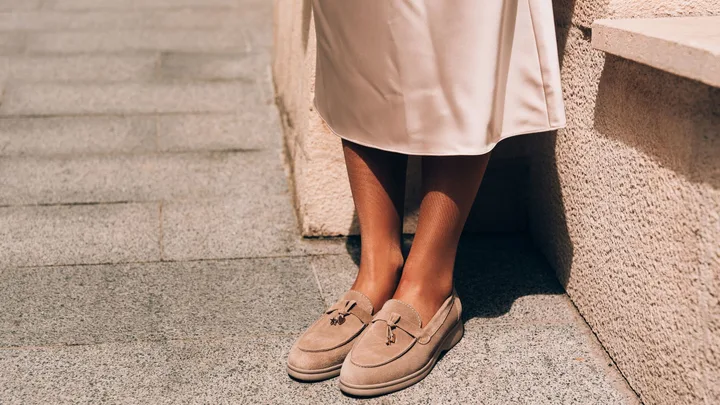 Close-up of a woman’s feet in light brown suede loafers with decorative gold charms and tassels. She is wearing a cream-coloured midi skirt against a sunlit stone wall.