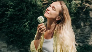 A woman with long blonde hair stands outdoors in the sunlight, holding a small, mint green portable handheld fan up to her face. She has her eyes closed and a relaxed expression, enjoying the breeze. The background features lush green foliage and a stone wall.