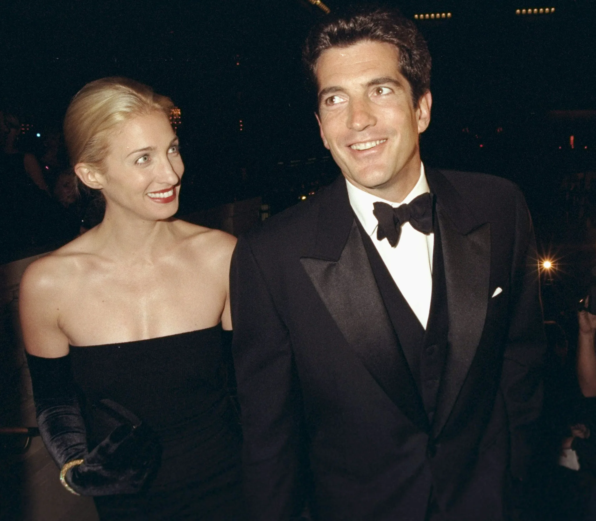 Carolyn wears a black strapless evening gown and her hair swept up behind her. JFK jr waits black tie. They smile at eachother.