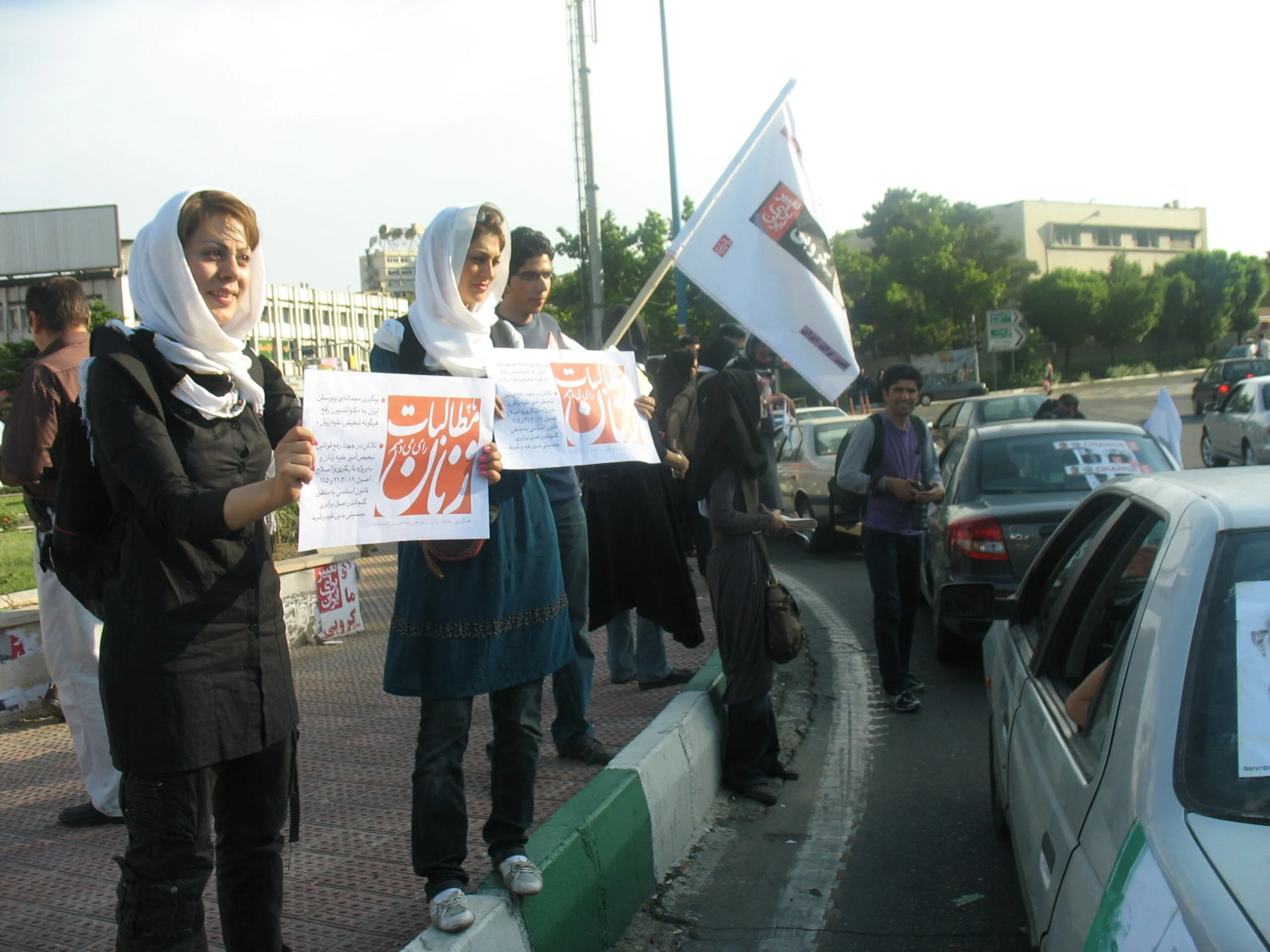 A group protests by the side of the road in Tehran. They carry flags and banners.