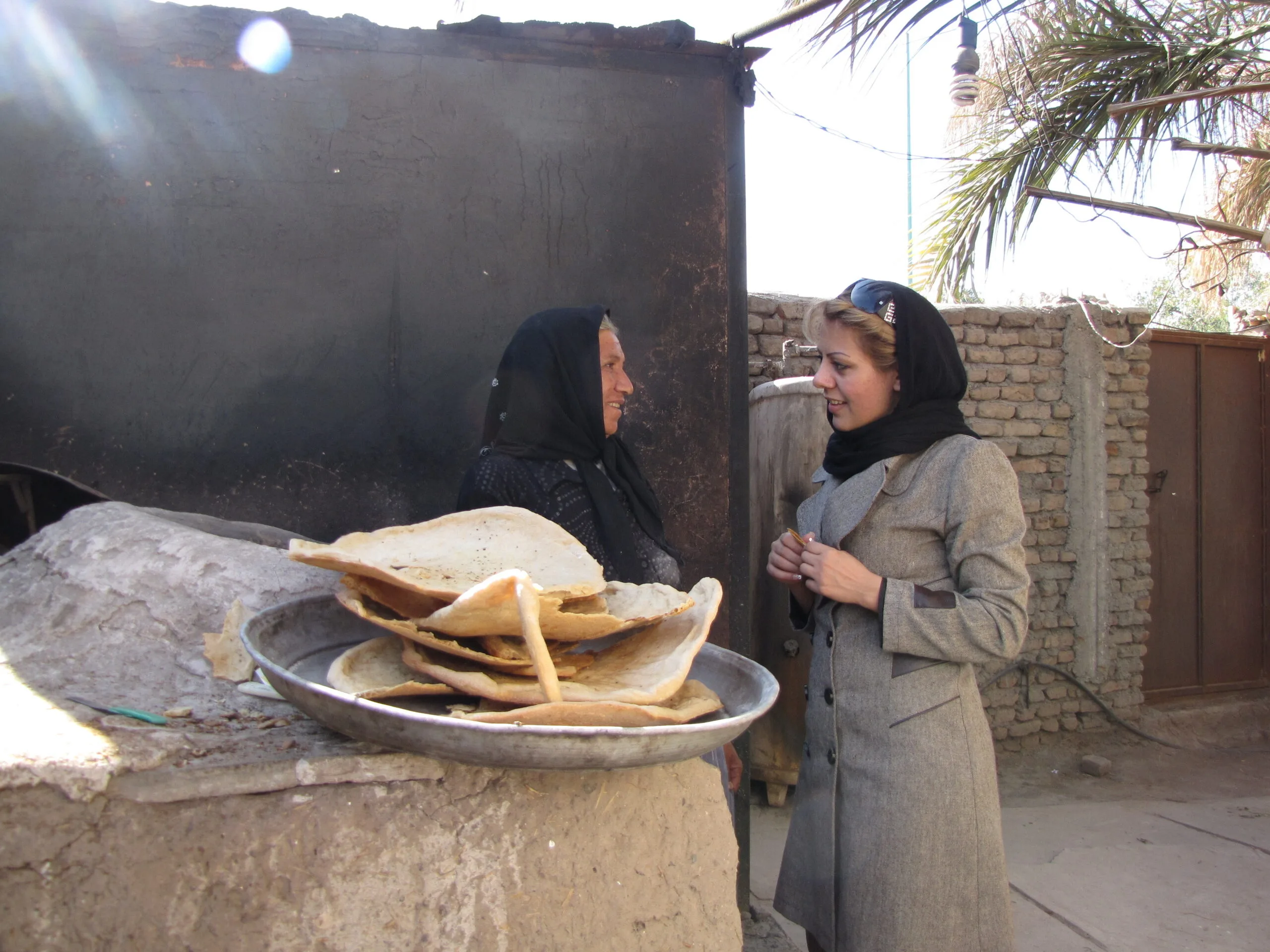 Saba in a black headscarf and tweed coat talks to a baker at her stall which is piled with flatbread.