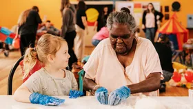 A young girl and an older Aboriginal woman sit together at a table making damper.