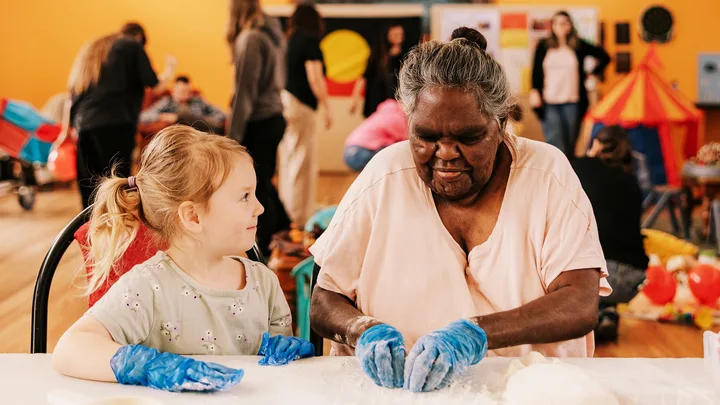 A young girl and an older Aboriginal woman sit together at a table making damper.