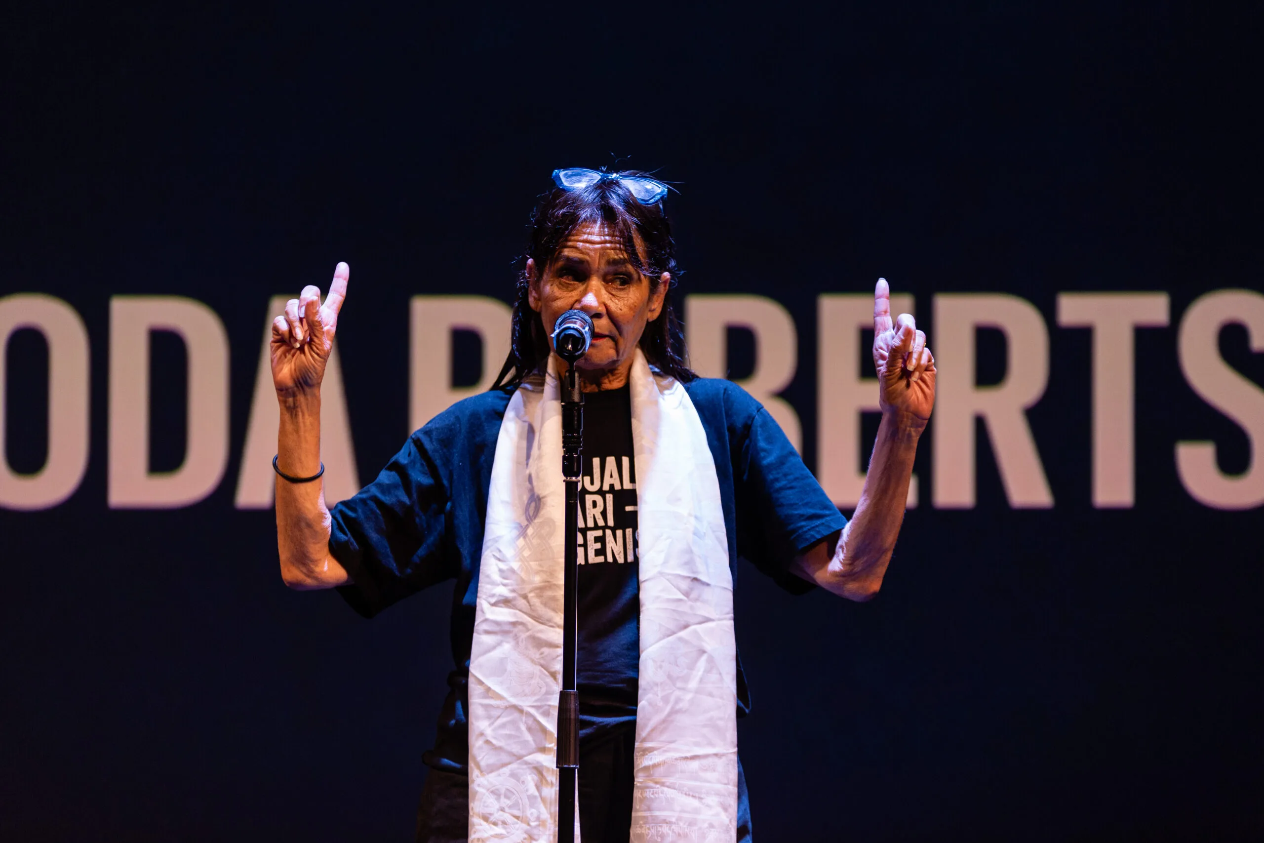 Rhoda Roberts wears a navy printed T-Shirt and a white scarf. She is pointing with both hands skyward.