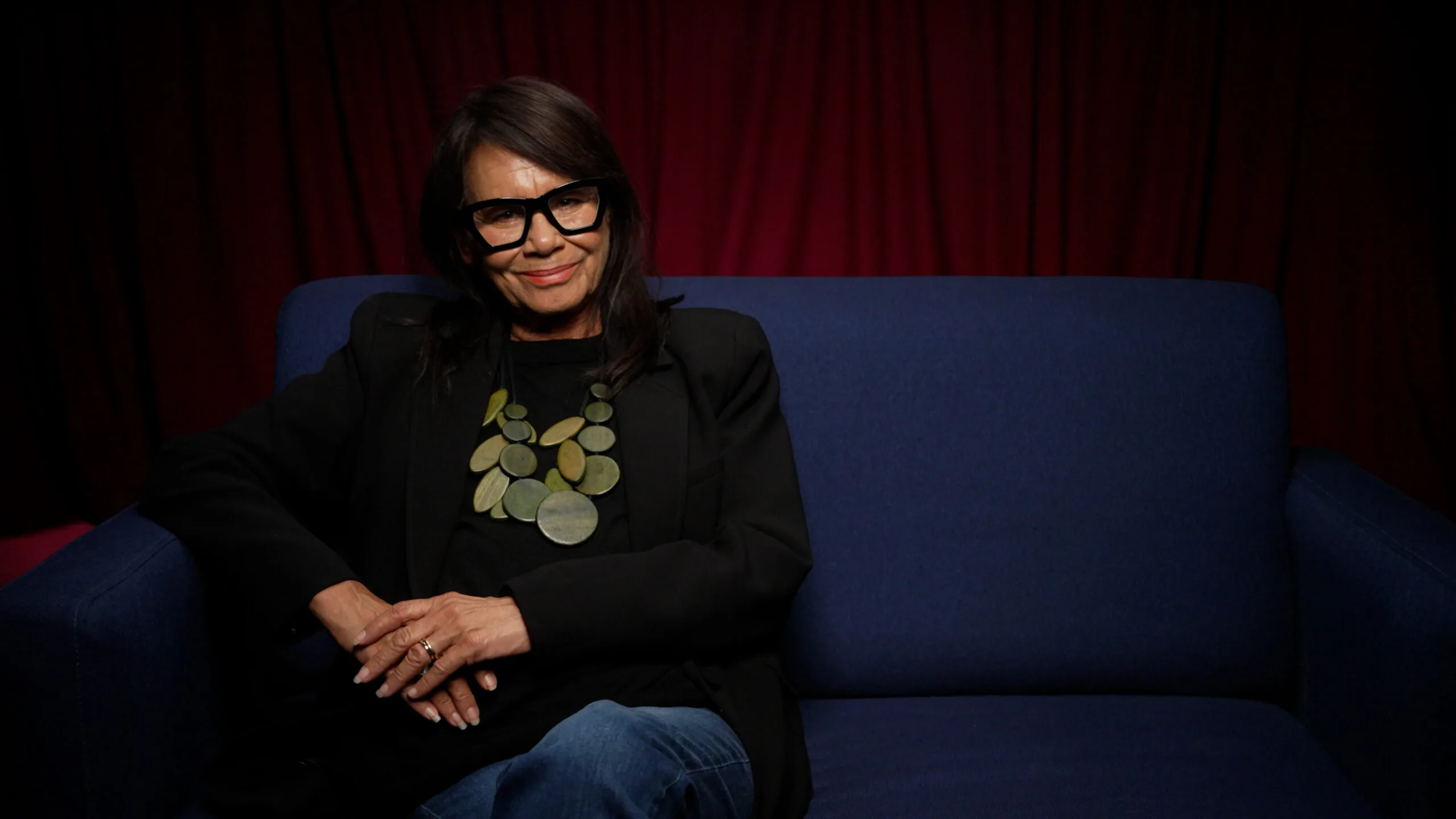 Rhoda Roberts sits ona  couch wearing jeans and a black jumper, with black rimmed glasses and chunky jewellery in shades of eucalypt gren.