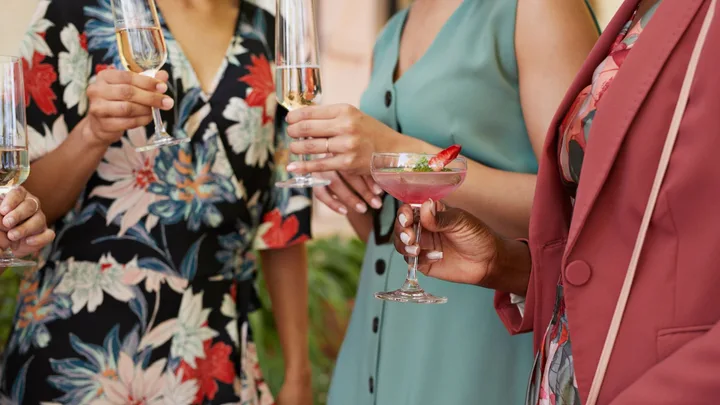 Three women laughing at an outdoor garden wedding wearing colourful midi-length wedding guest dresses and holding cocktail glasses.