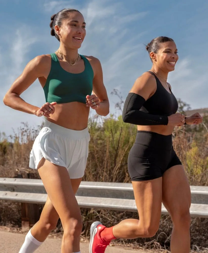 Two women, Lululemon ambassadors Kayla Jeter and Montana Farrah-Seaton, smiling while running along an outdoor path. They are wearing athletic gear, including the Go Further sports bra, shorts and running shoes.