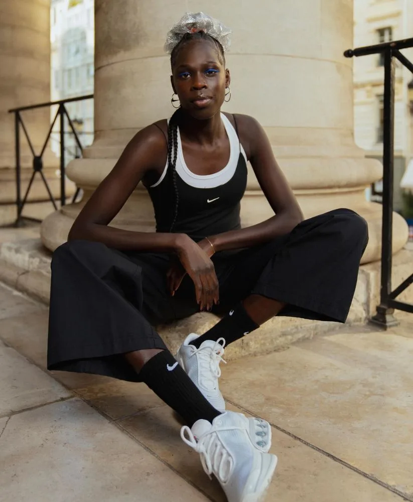 A woman sitting cross-legged on a stone ledge in front of a large architectural column. She is wearing a black Nike tank top layered over a white top, wide-leg black trousers, black Nike crew socks and chunky white sneakers. Her look is styled with blue eyeliner and a silver mesh hair accessory.