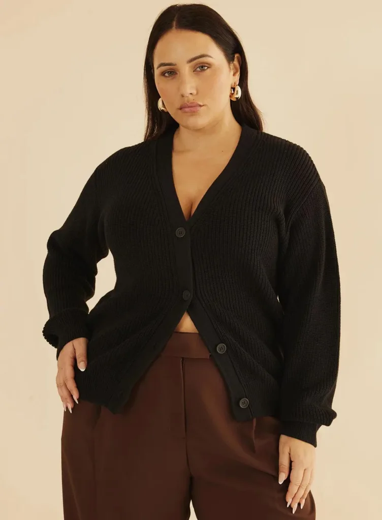 A model posing in an Atmos&Here Curvy black button-down cardigan with ribbed texture, paired with high-waisted brown tailored pants and gold hoop earrings.