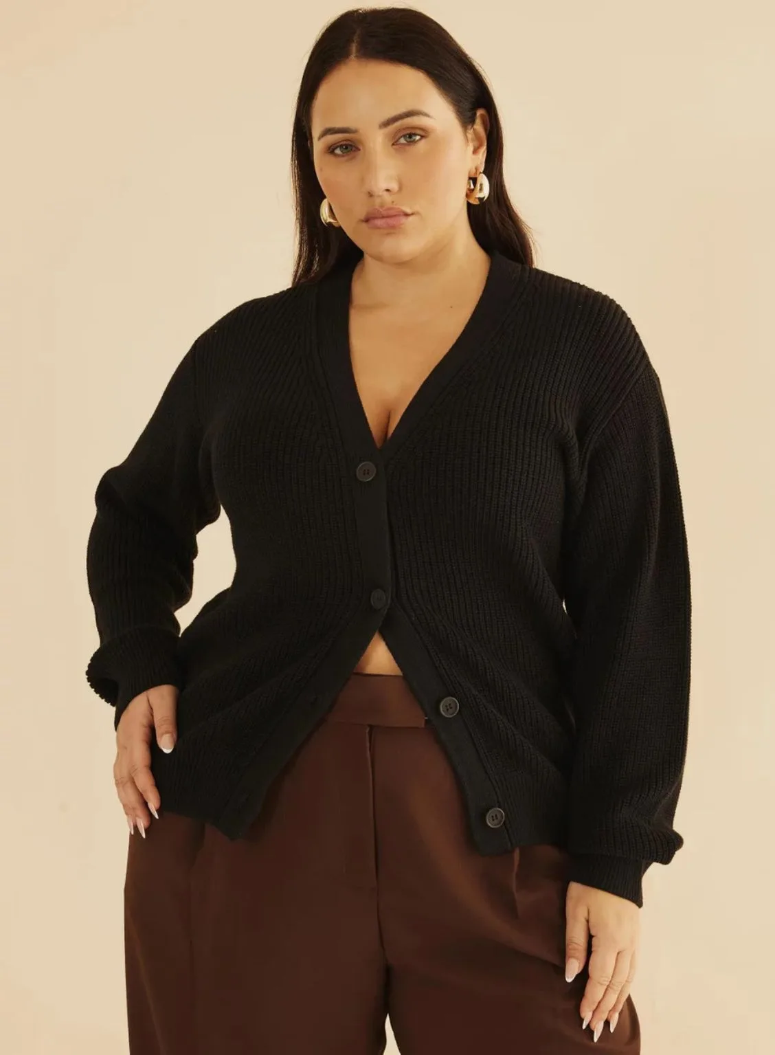 A model posing in an Atmos&Here Curvy black button-down cardigan with ribbed texture, paired with high-waisted brown tailored pants and gold hoop earrings.