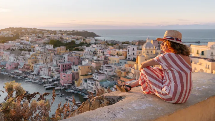 A woman in a striped dress and straw hat sits on a stone wall, looking out over a coloruful coastal European town and harbour during a golden hour sunset.