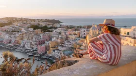 A woman in a striped dress and straw hat sits on a stone wall, looking out over a coloruful coastal European town and harbour during a golden hour sunset.