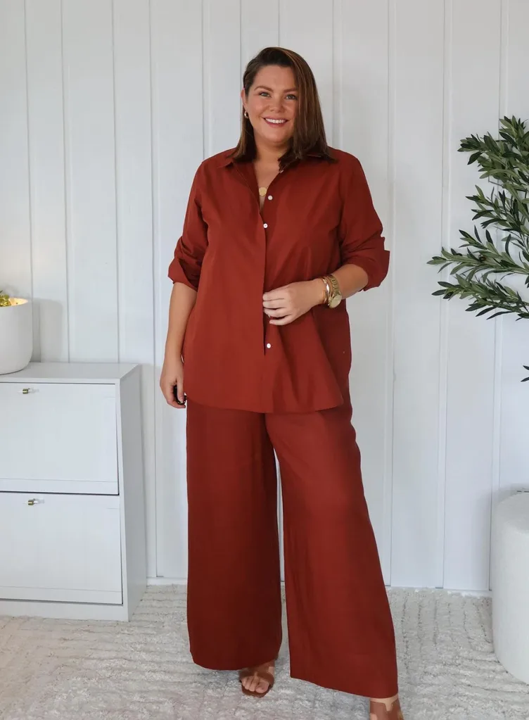 A woman smiling while wearing a matching rust-coloured button-down shirt and wide-leg trousers set.