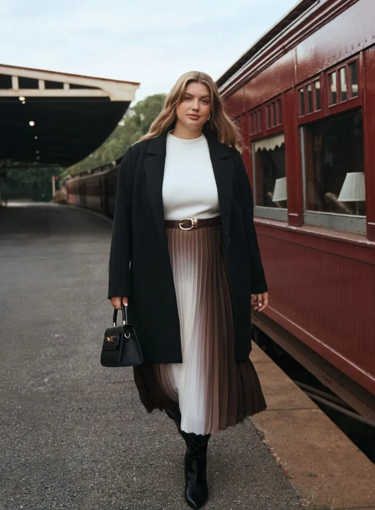 A woman walking along a vintage train platform wearing a black coat, white top, and a brown and white ombré pleated skirt.