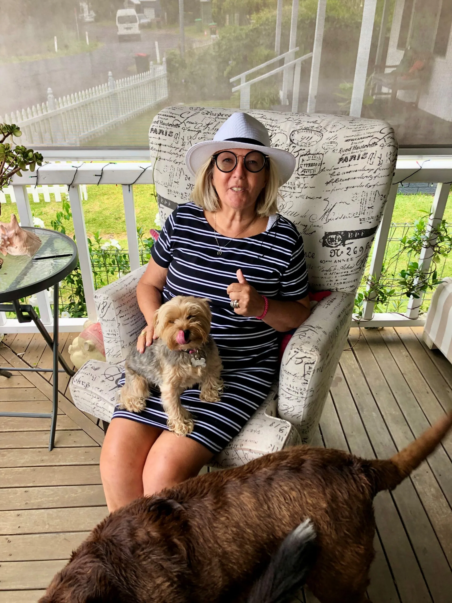 Mandy sits on the veranda with a dog on her lap and another at her feet. She is wearing a striped dress and a white hat.