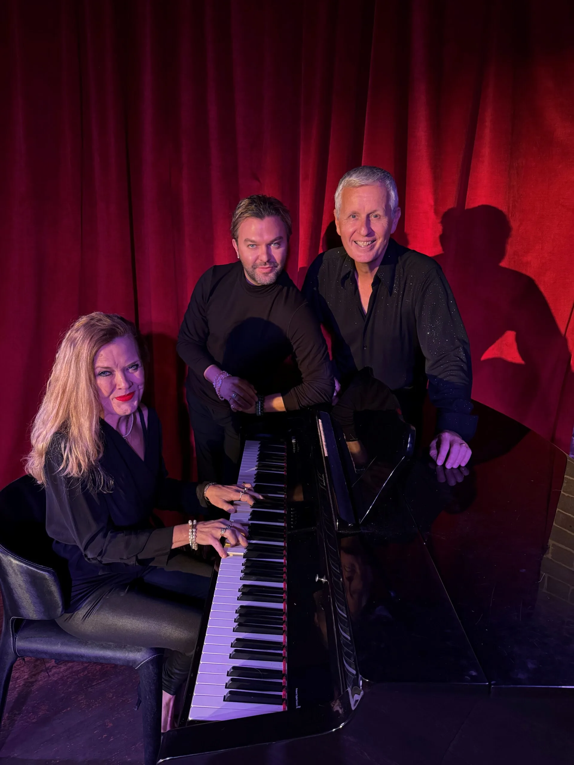 The three musicians stand around a piano in front of a red velvet curtain.