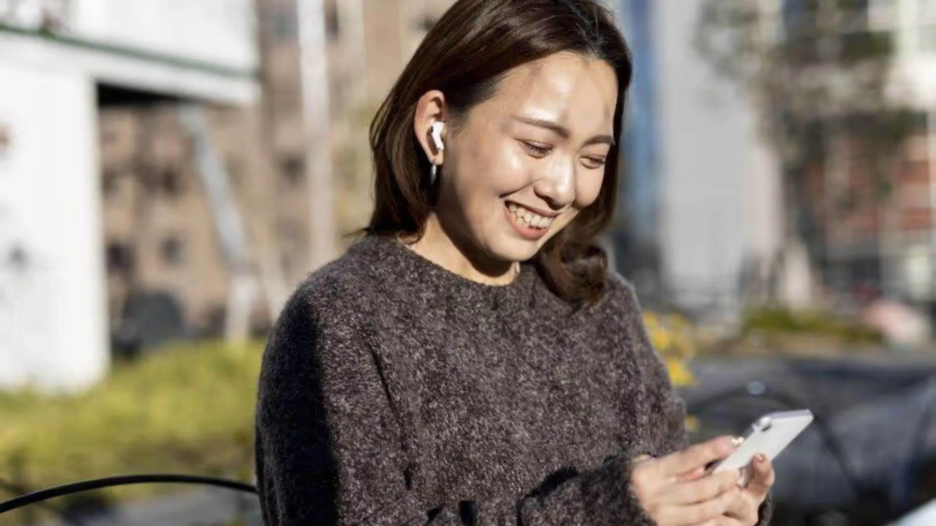 A smiling woman wearing wireless earbuds and looking down at her smartphone outdoors on a sunny day.